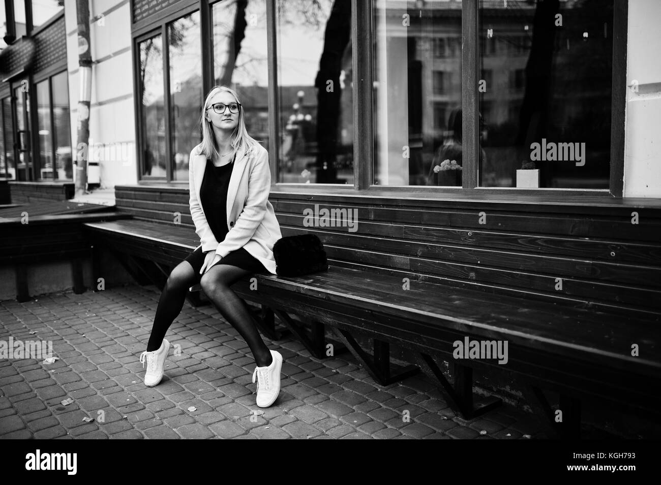 Blonde girl at glasses and pink coat, black tunic sitting on bench at street. Stock Photo