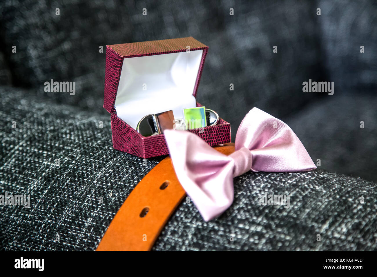 The groom's sophisticated accessories including cuff links in the box, leather brown belt and pink bow tie, placed on a grey sofa Stock Photo