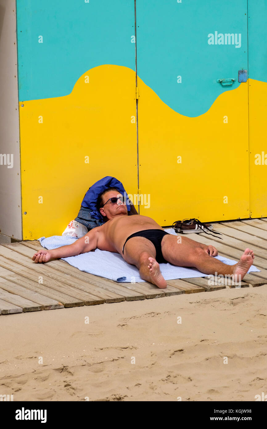 Man wearing small swimming trunks sunbathing behind beach huts Dunkirk Stock Photo