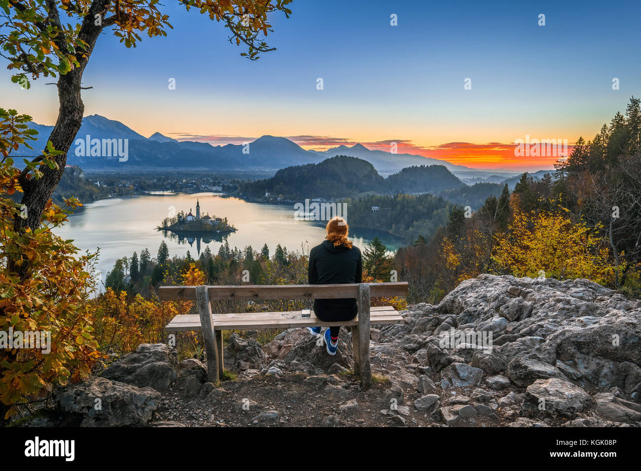 Bled, Slovenia - Red hair runner woman relaxing and enjoying the beautiful autumn view and the colorful sunrise of Lake Bled sitting on a hilltop benc Stock Photo
