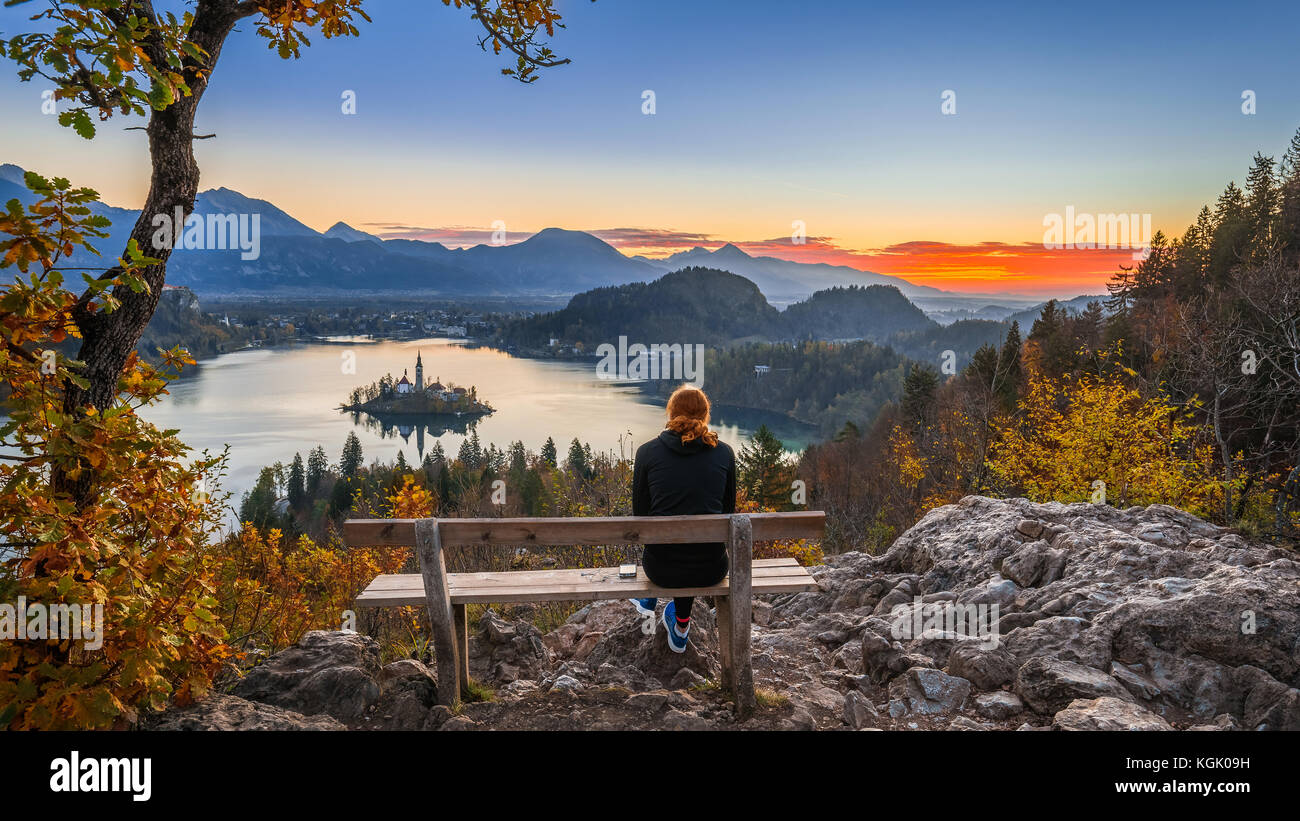 Bled, Slovenia - Red hair runner woman relaxing and enjoying the beautiful autumn view and the colorful sunrise of Lake Bled sitting on a hilltop benc Stock Photo