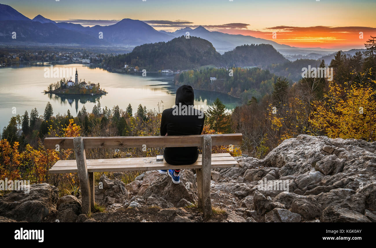 Bled, Slovenia - Runner woman relaxing and enjoying the beautiful autumn view and the colorful sunrise of Lake Bled sitting on a hilltop bench wearing Stock Photo