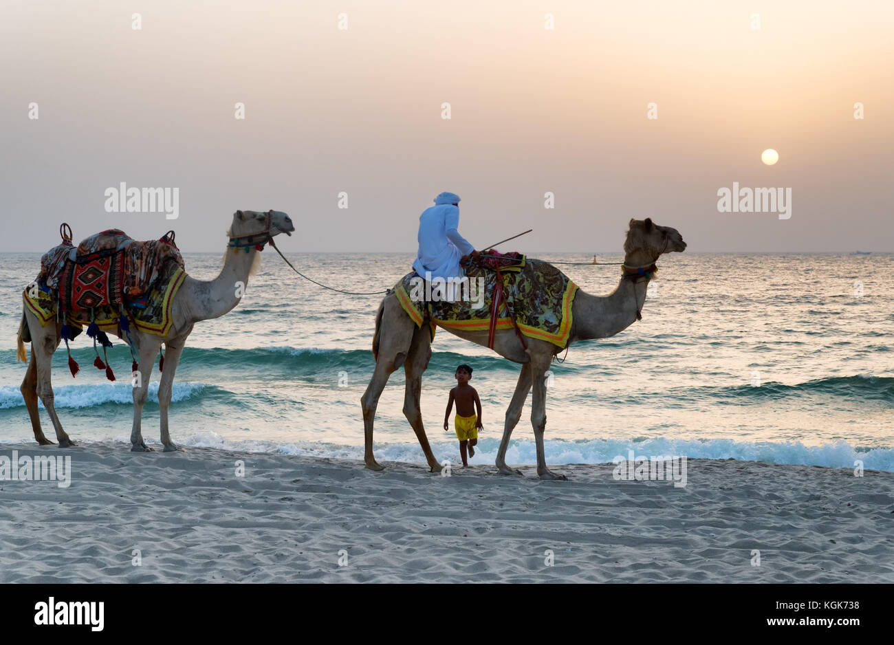Boy stares up in amazement at Arabic man wearing a traditional thawb riding a camel in Jumeriah Beach Resort, Dubai, United Arab Emirates Stock Photo