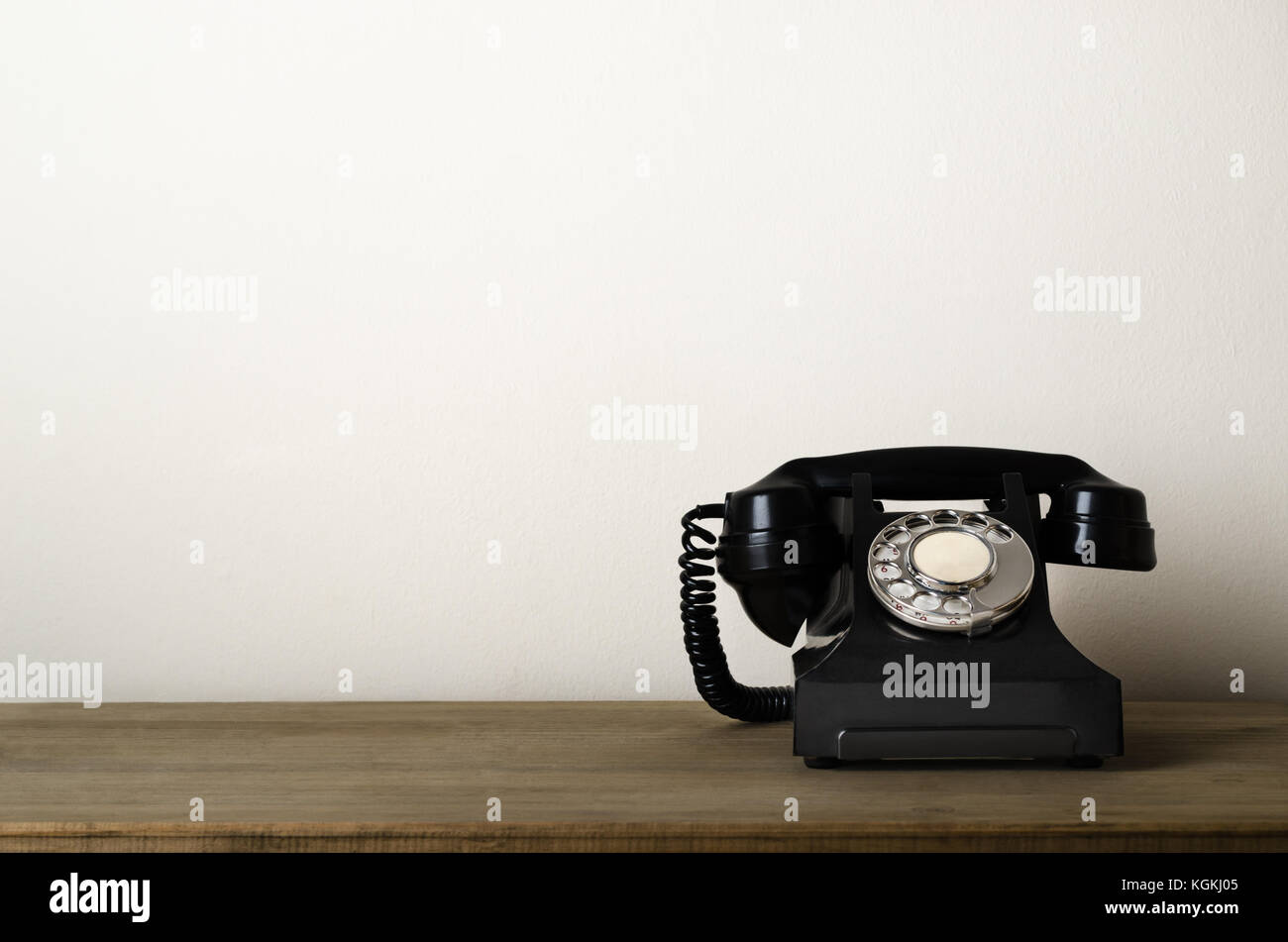 Black, 1940s vintage bakelite telephone at eye level on a wooden desk with ivory white wall as background providing copy space to the left. Stock Photo