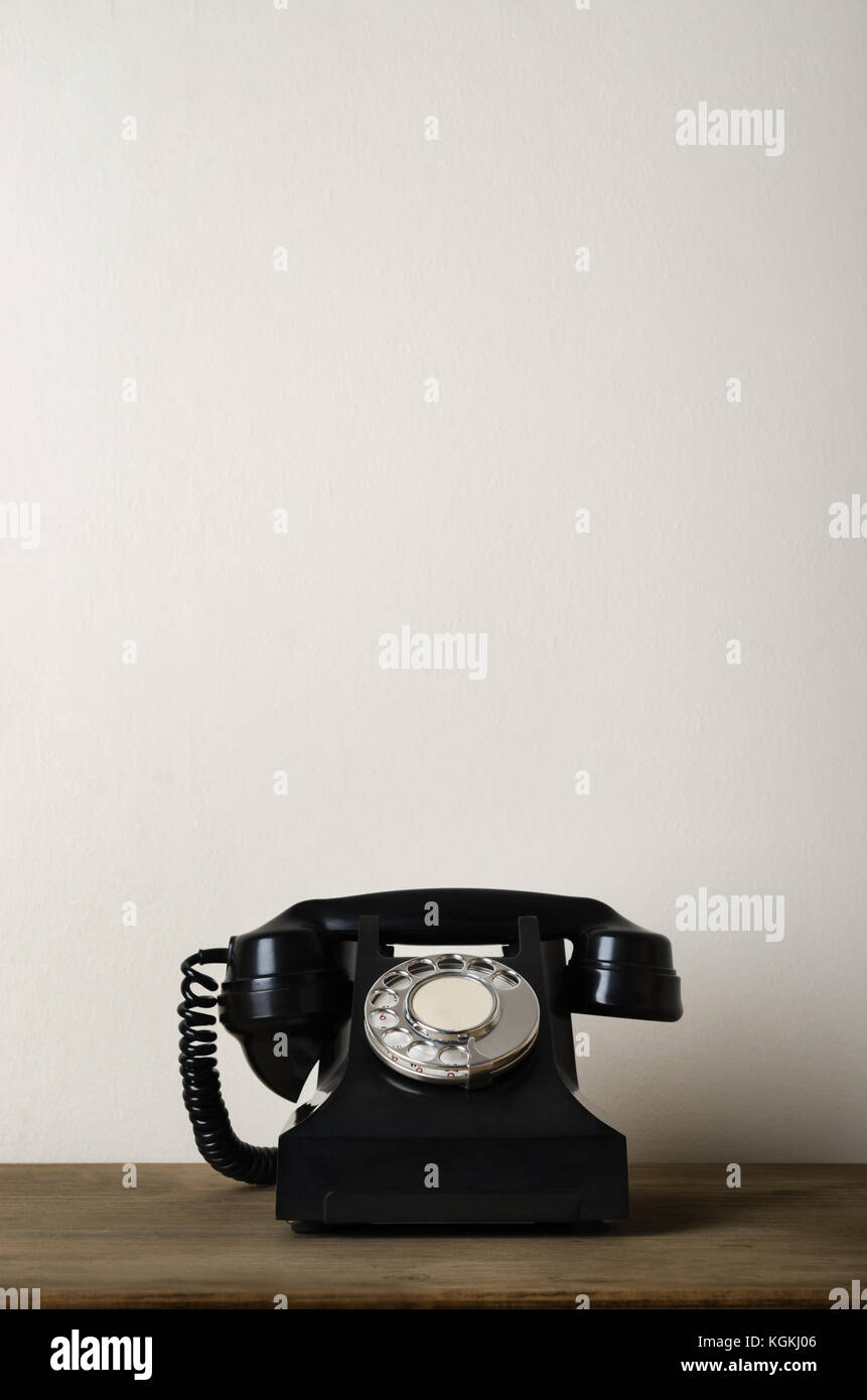 Black 1940s vintage bakelite telephone on a wooden desk with ivory white wall as background providing copy space above. Stock Photo