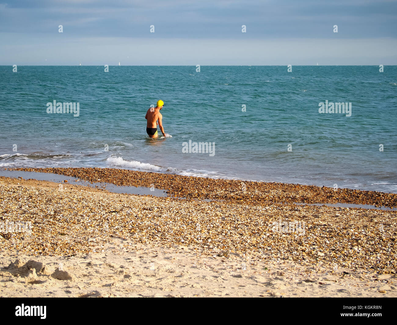 A man wearing swimming trunks and swim hat wades into the solent in an Autmn day Stock Photo