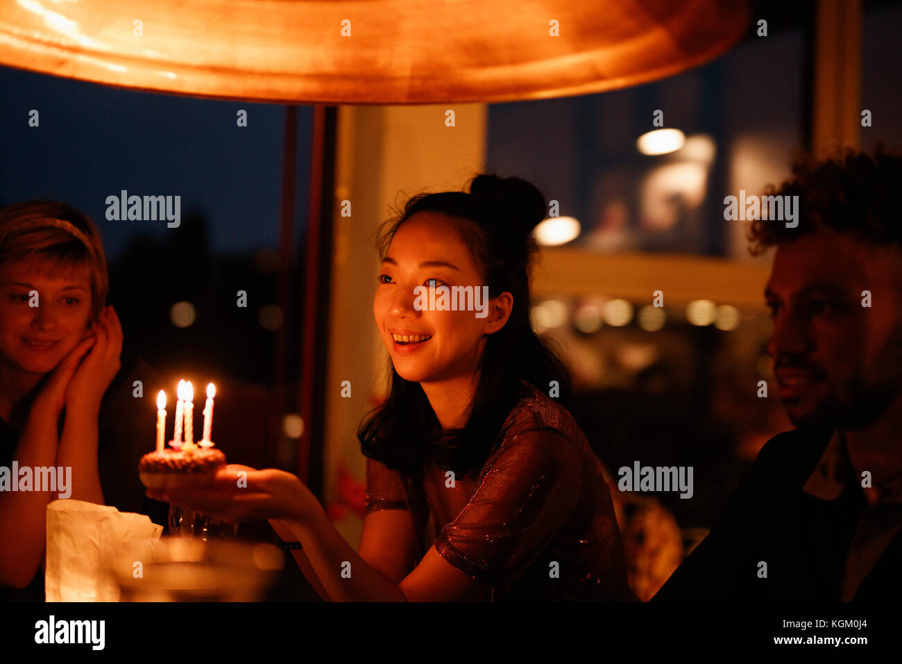 Happy woman holding small birthday cake with lit candles while sitting by friends at home Stock Photo
