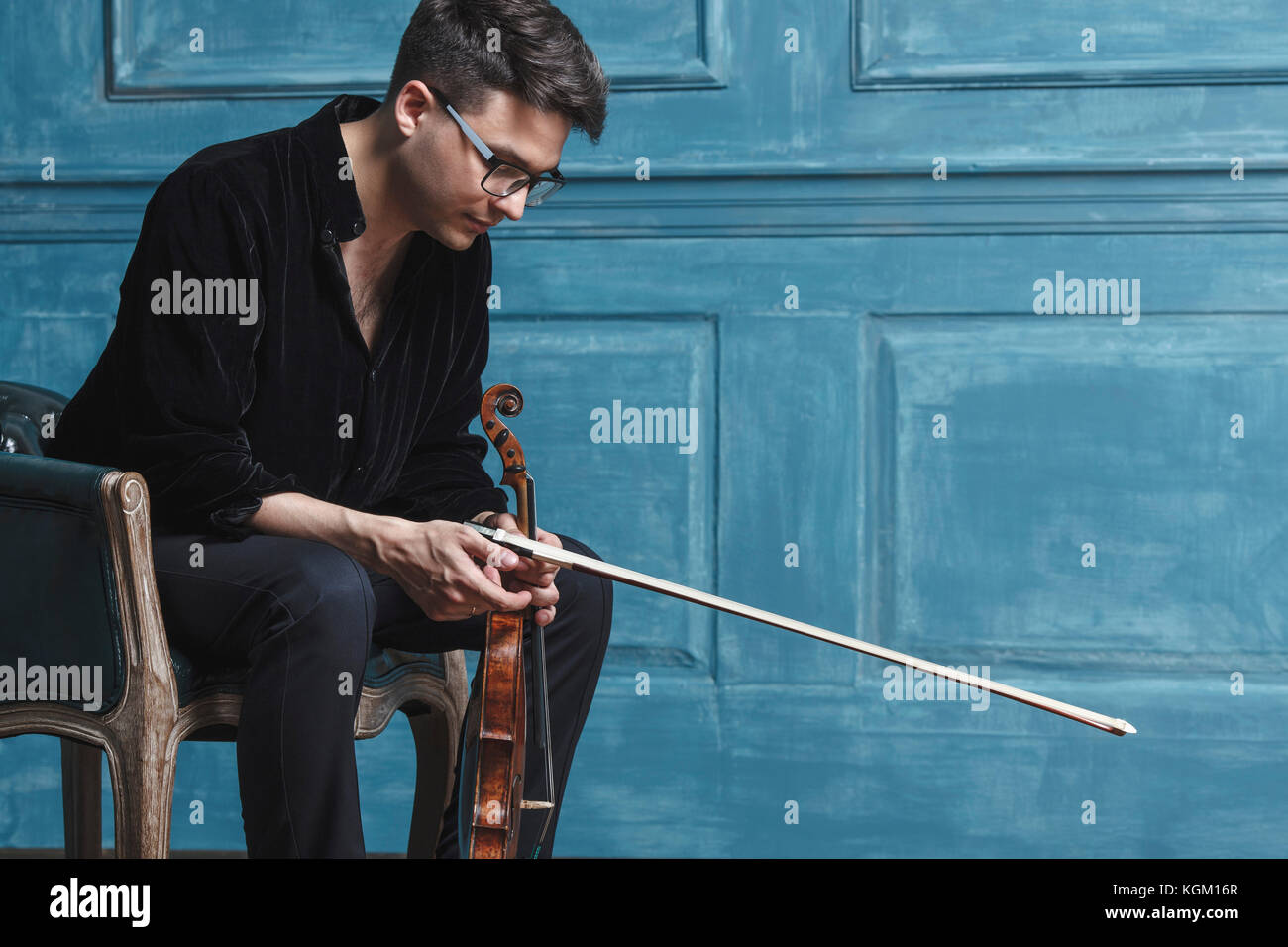 Young man holding violin while sitting on chair by blue wall Stock Photo