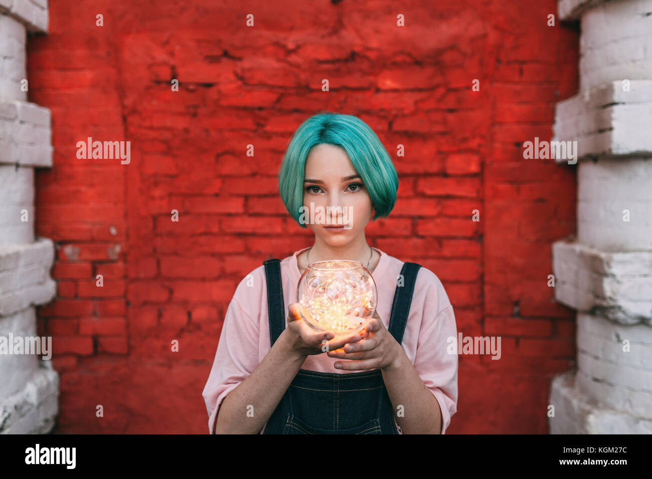 Portrait of teenage girl holding illuminated string lights while standing against brick wall Stock Photo
