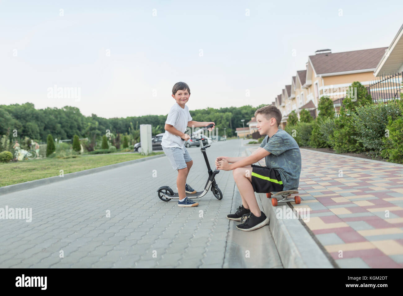 Boy sitting on skateboard while brother riding push scooter on cobbled street Stock Photo