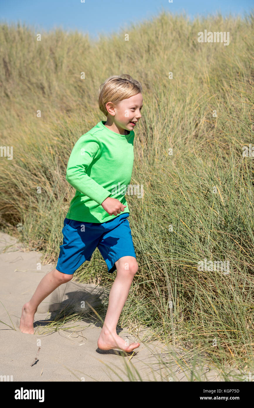 Young blond boy running barefoot on sand with sea grass, wearing blue shorts, green shirt, vertical portrait of child at beach. Stock Photo