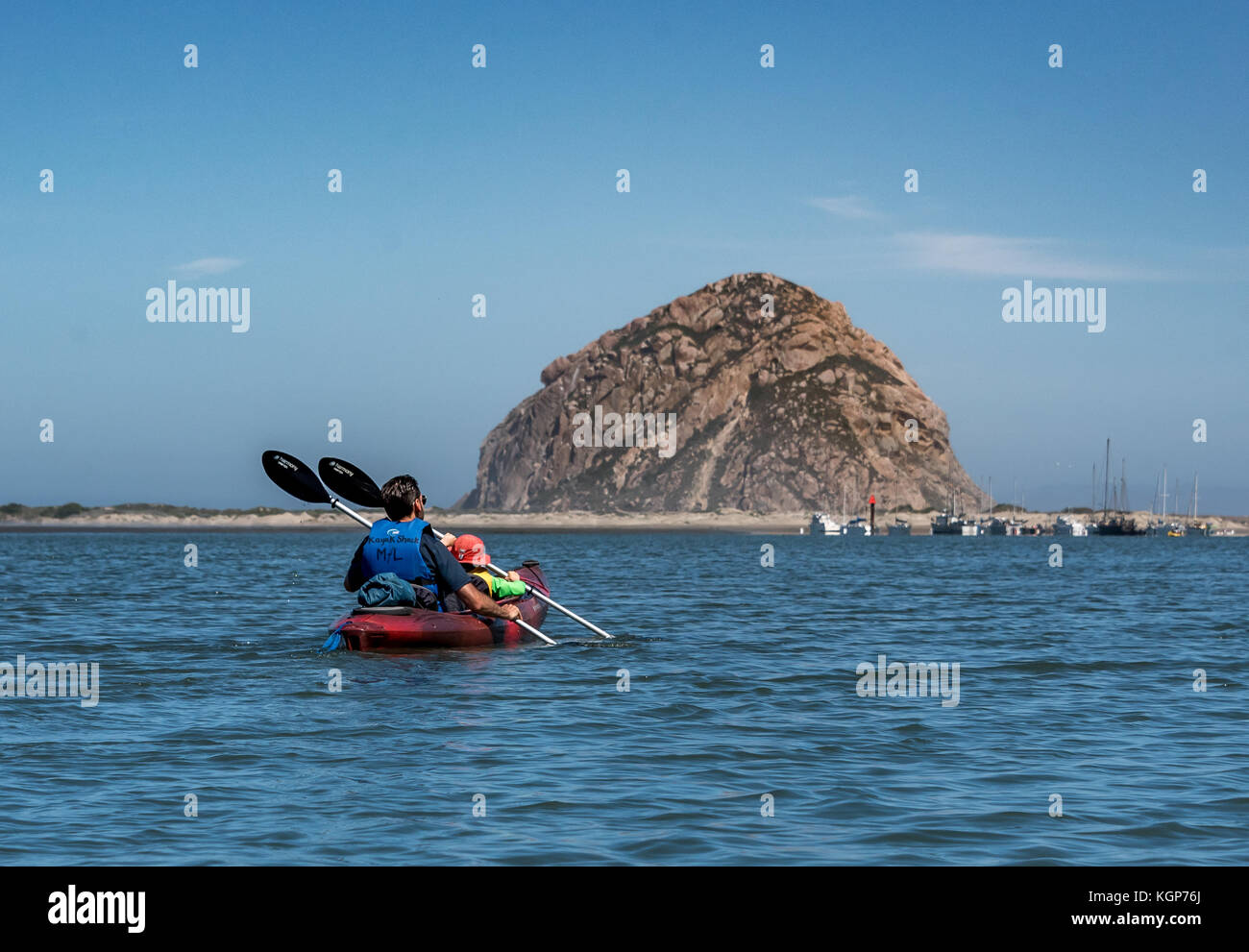 A father and son paddle a tandem red kayak toward Morro Rock on Morro Bay, the young boy wears an orange sun hat, both are paddling together Stock Photo