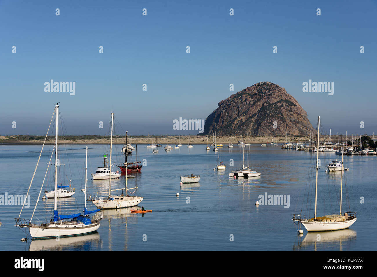 View of Morro Rock across Morro Bay with sail boats anchored across flat water, an orange kayak and kayaker pass between the boats on a clam morning. Stock Photo