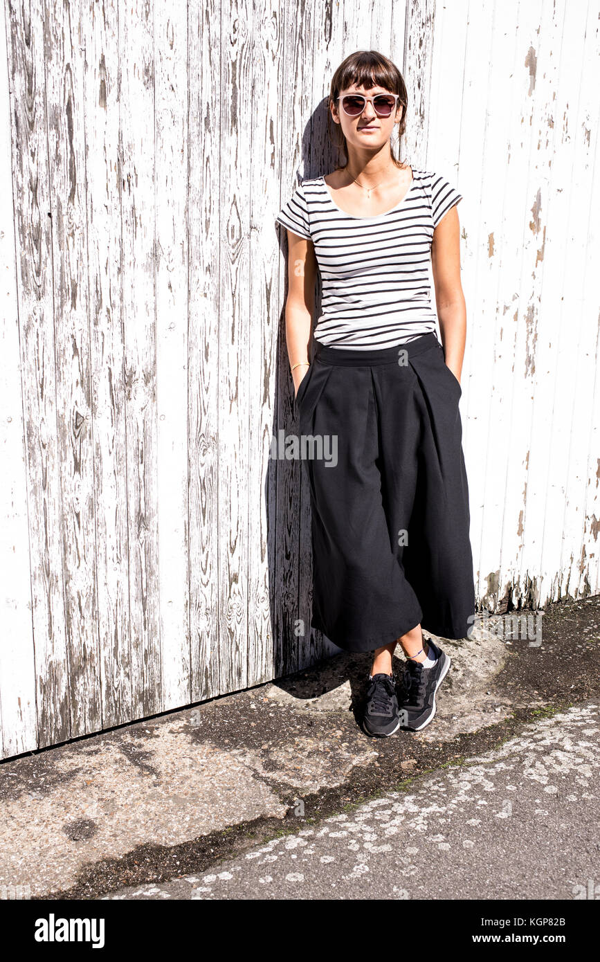Hipster woman wearing sunglasses dressed in urban minimal style with black striped T-shirt, wide leg style black trousers and sneakers. Shot on a East Stock Photo