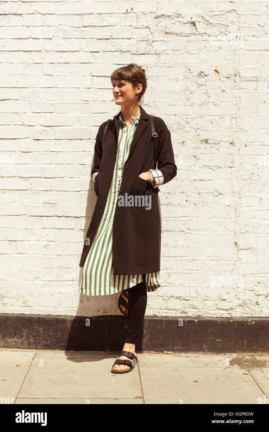 Hipster woman dressed in urban minimal style with long black jacket, striped dress, black trousers and sandals. Shot on a street with white brick wall Stock Photo