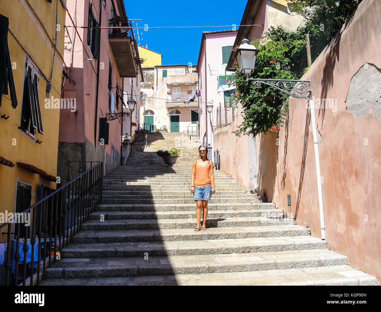 Young woman with sunglasses dressed in casual summer clothes walking down an alley in a small rural medieval village in Italy Stock Photo