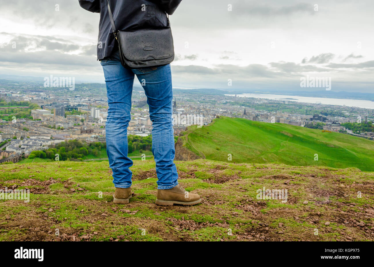 Young female model wearing skinny blue jeans and brown suede boots admiring Edinburgh landscape from the top of the Arthur's seat. Scotland, UK. Stock Photo