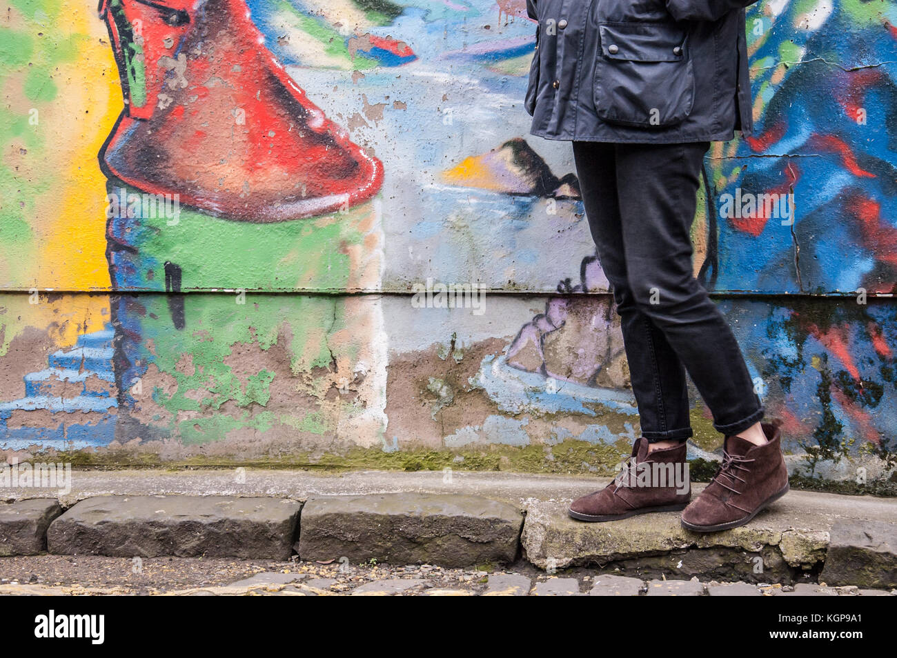Model wearing tight black denim skinny trousers and brown suede ankle boots and walking in front of a graffiti wall. Focus on legs and shoes. Stock Photo