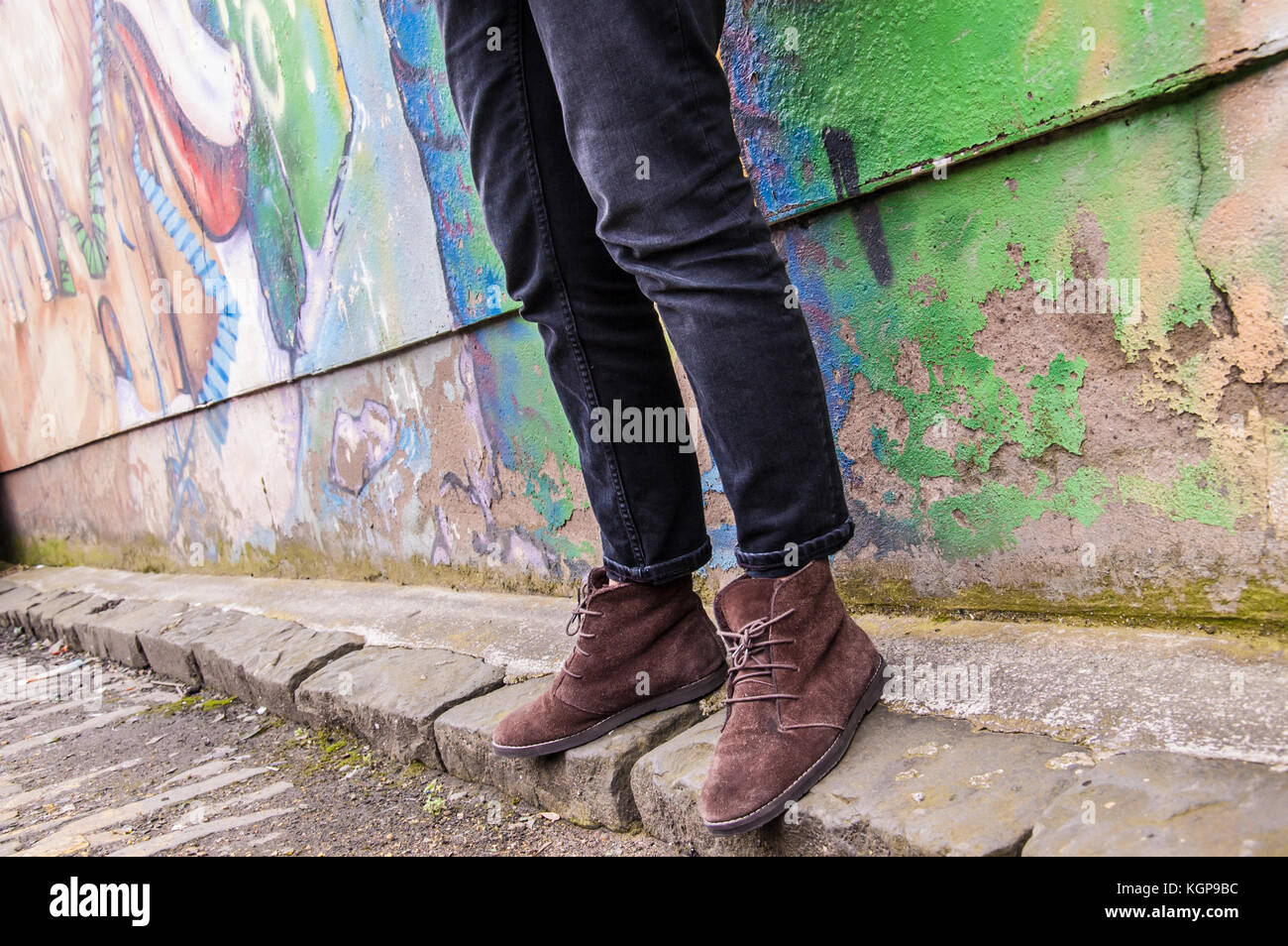 Model wearing tight black denim skinny trousers and brown suede ankle boots and walking in front of a graffiti wall. Focus on legs and shoes. Surface  Stock Photo