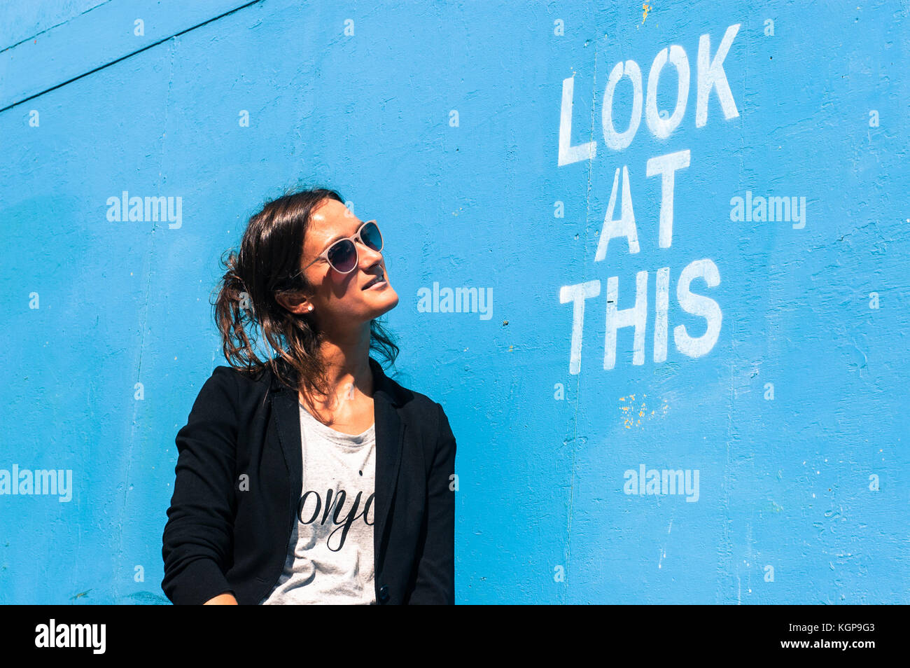 Hipster model wearing sunglasses posing next to a blue wall with the words 'Look at this' written on it Stock Photo