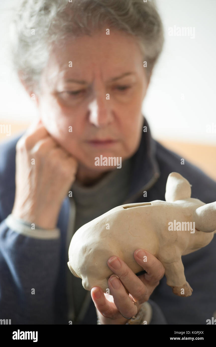 Older woman saver with money piggy bank, looking depressed, with head leaning on hand, savings account interest rates remain all time low, 2017. Stock Photo