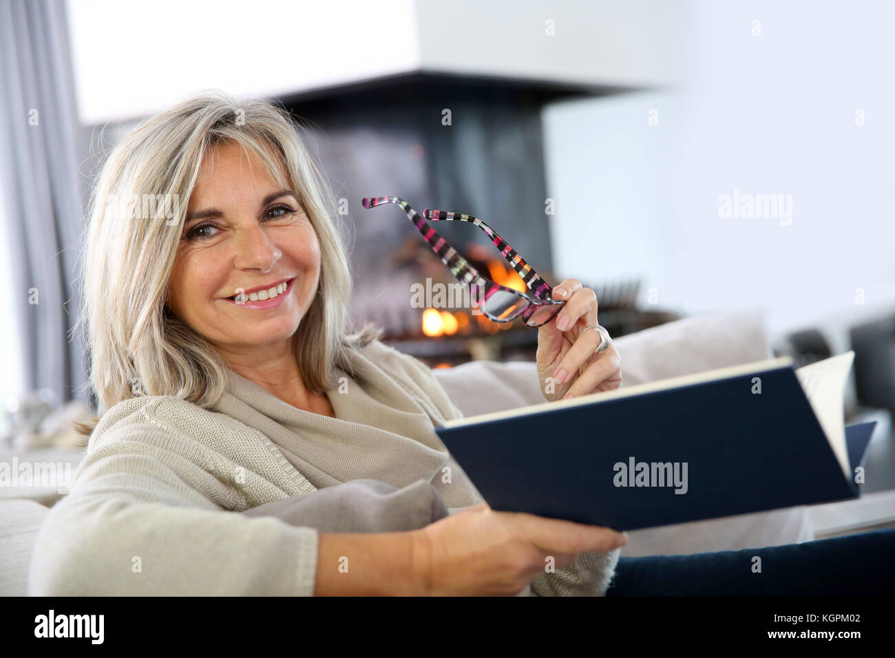 Senior woman reading book at home by fireplace Stock Photo