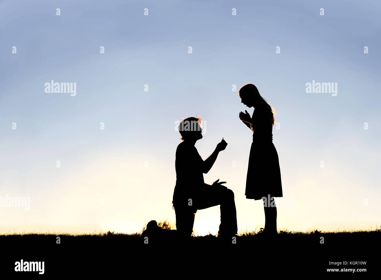 A silhouette of a young man, down on one knee and holding a diamond engagement ring, proposing to his girlfriend. Stock Photo