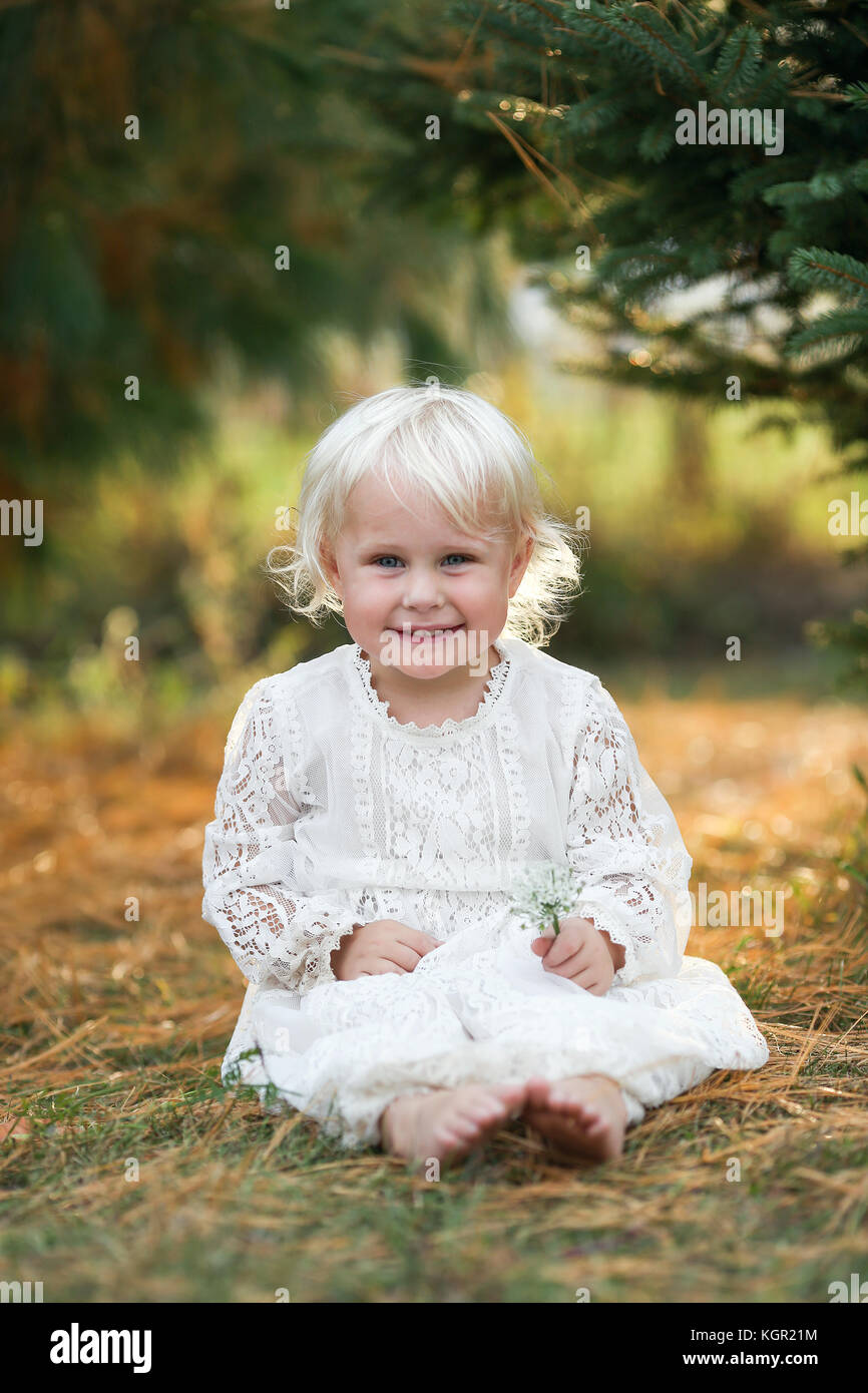 A beautiful 2 year old girl child is sitting outside, smiling under the pine trees at sunset on a late summer evening. Stock Photo