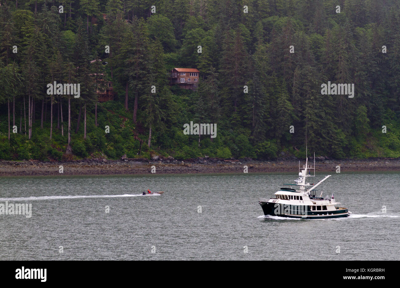Two men in an outboard motorboat passing a fishing boat along the Alaskan coast near Ketchikan with secluded houses in forest in the background. Stock Photo