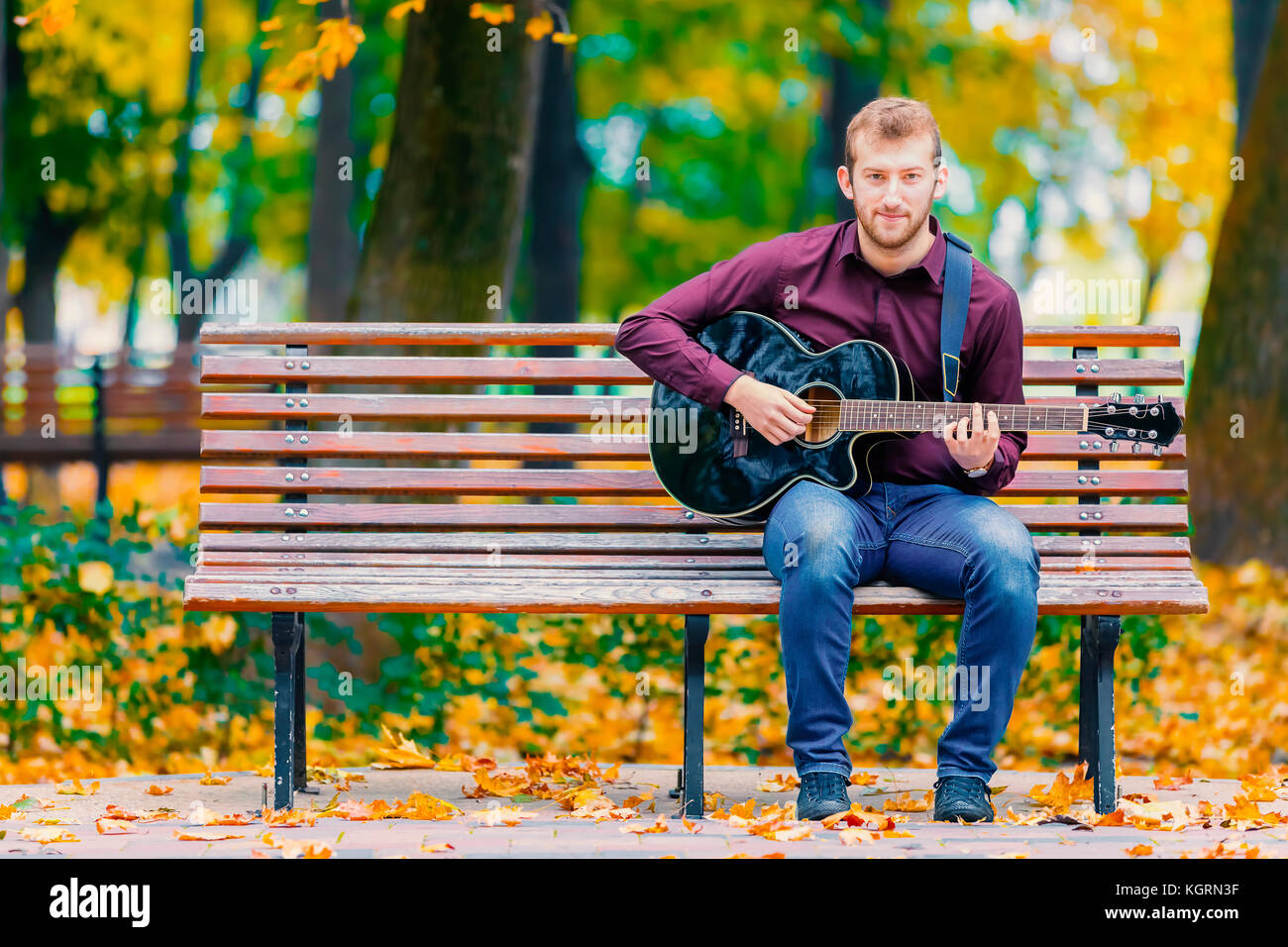 Young man sitting on bench and playing acoustic guitar in city park Stock Photo