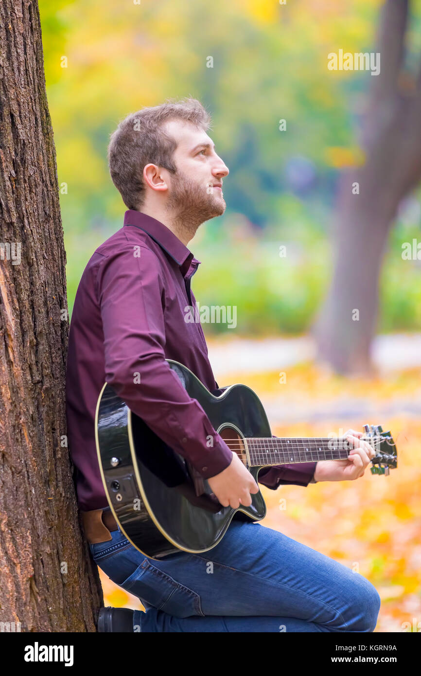 Young man sitting and playing acoustic guitar in city park. Focus on his eye! Stock Photo
