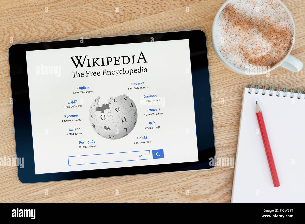 The Wikipedia website features on an iPad tablet device which rests on a wooden table beside a notepad and pencil and a cup of coffee (Editorial use o Stock Photo