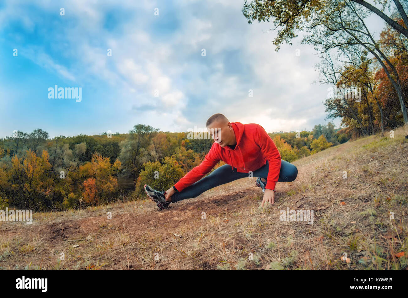 Slim caucasian jogger man in red jacket and black sports leggins performs warming-up  before jogging on the autumnal forest hil with sunset dramatic s Stock Photo