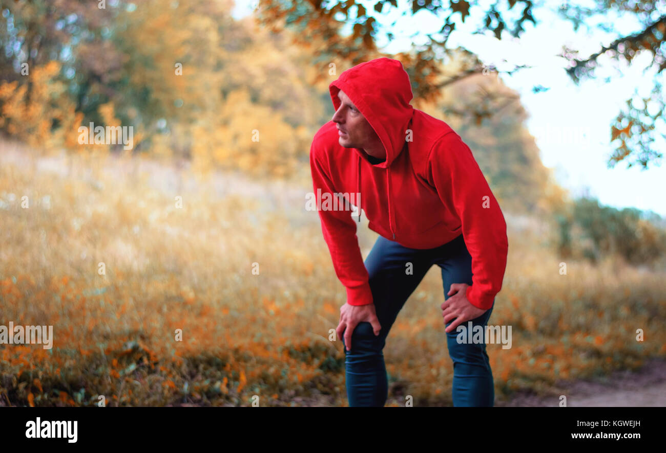A young jogger man in a red sports jacket with a hood and black sport leggins  rest after running on a bright yellow autumnall background. Stock Photo