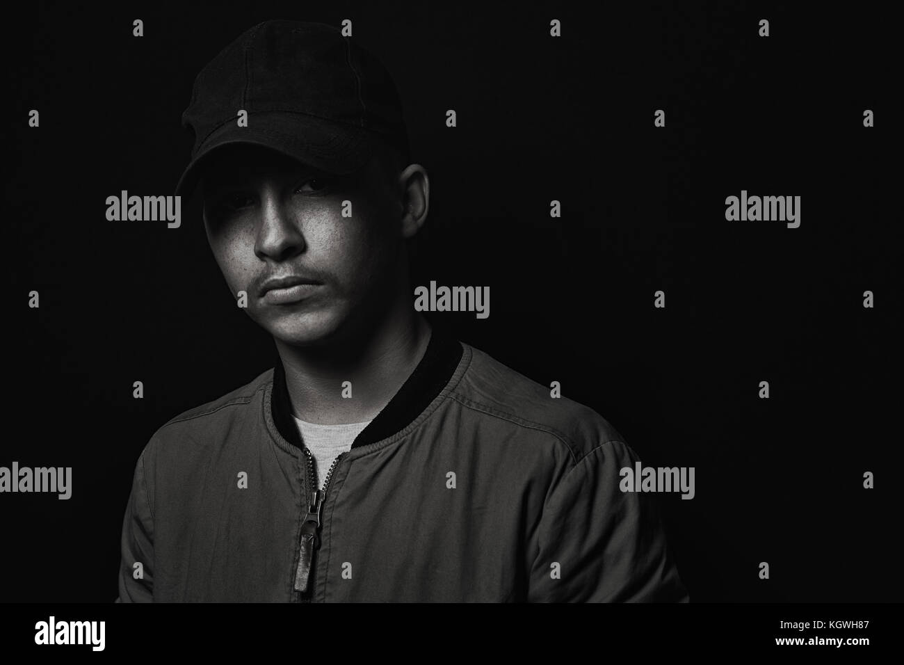 teen boy posing with black cap and bomber jacket in front of black background Stock Photo