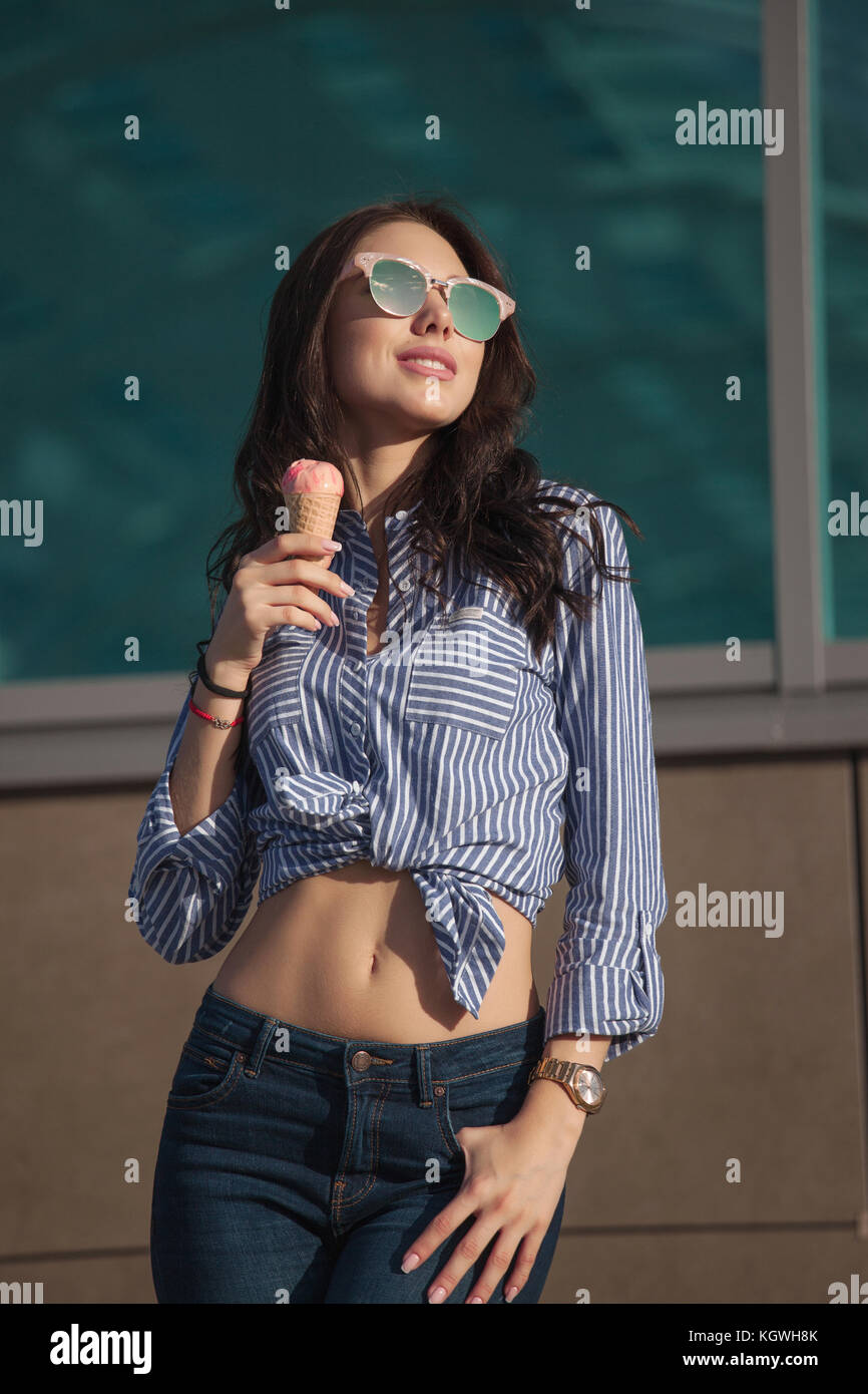 Young, beautiful brunette woman eating a pink ice cream in a waffle horn. She's in a good mood She wears glasses with dark glasses, a shirt in a white Stock Photo