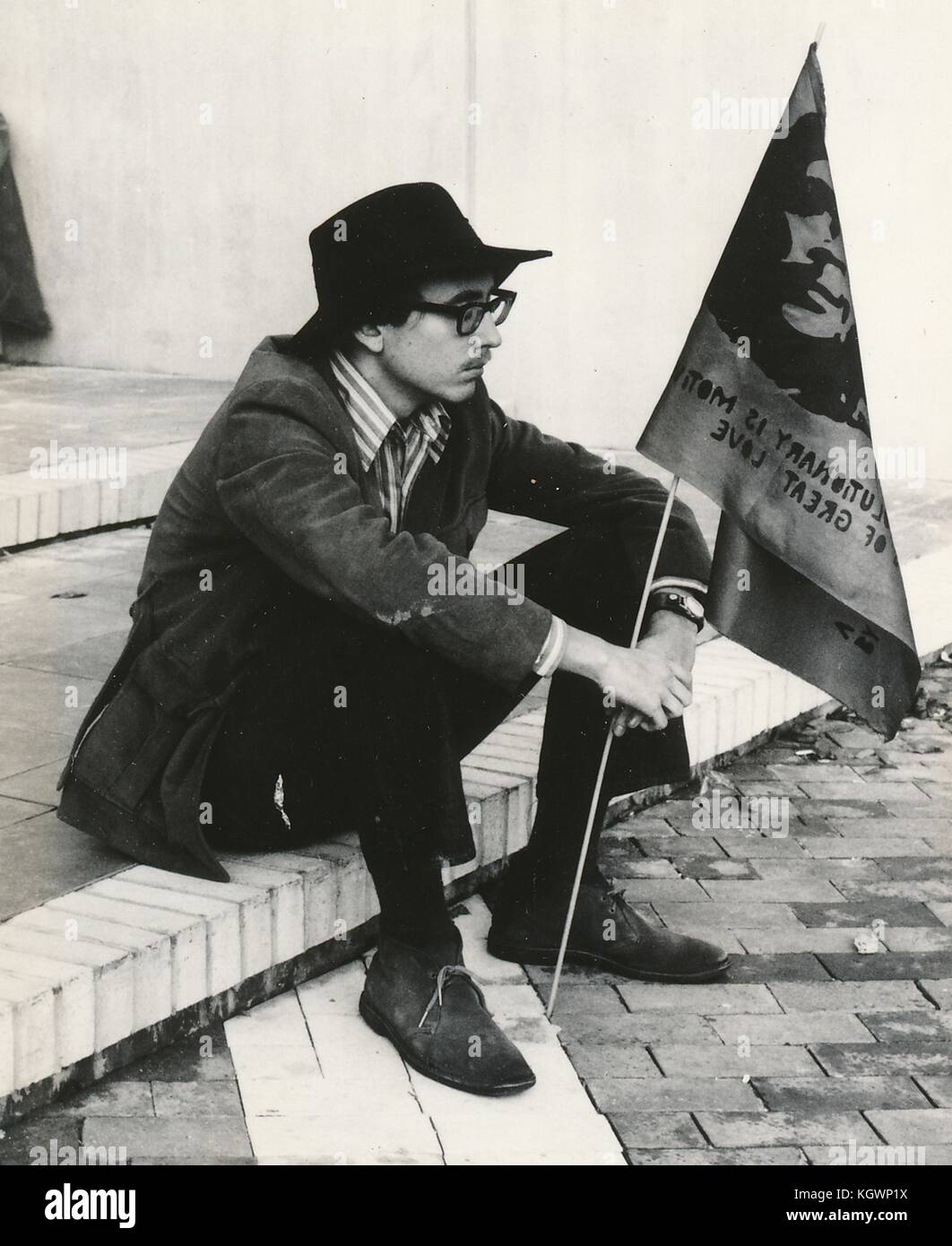 A male student wearing hippie attire, including thick glasses and a fedora hat, sits on a step and holds a flag with the face of revolutionary Che Guevara during an anti Vietnam War student sit-in protest at North Carolina State University, Raleigh, North Carolina, 1970. () Stock Photo