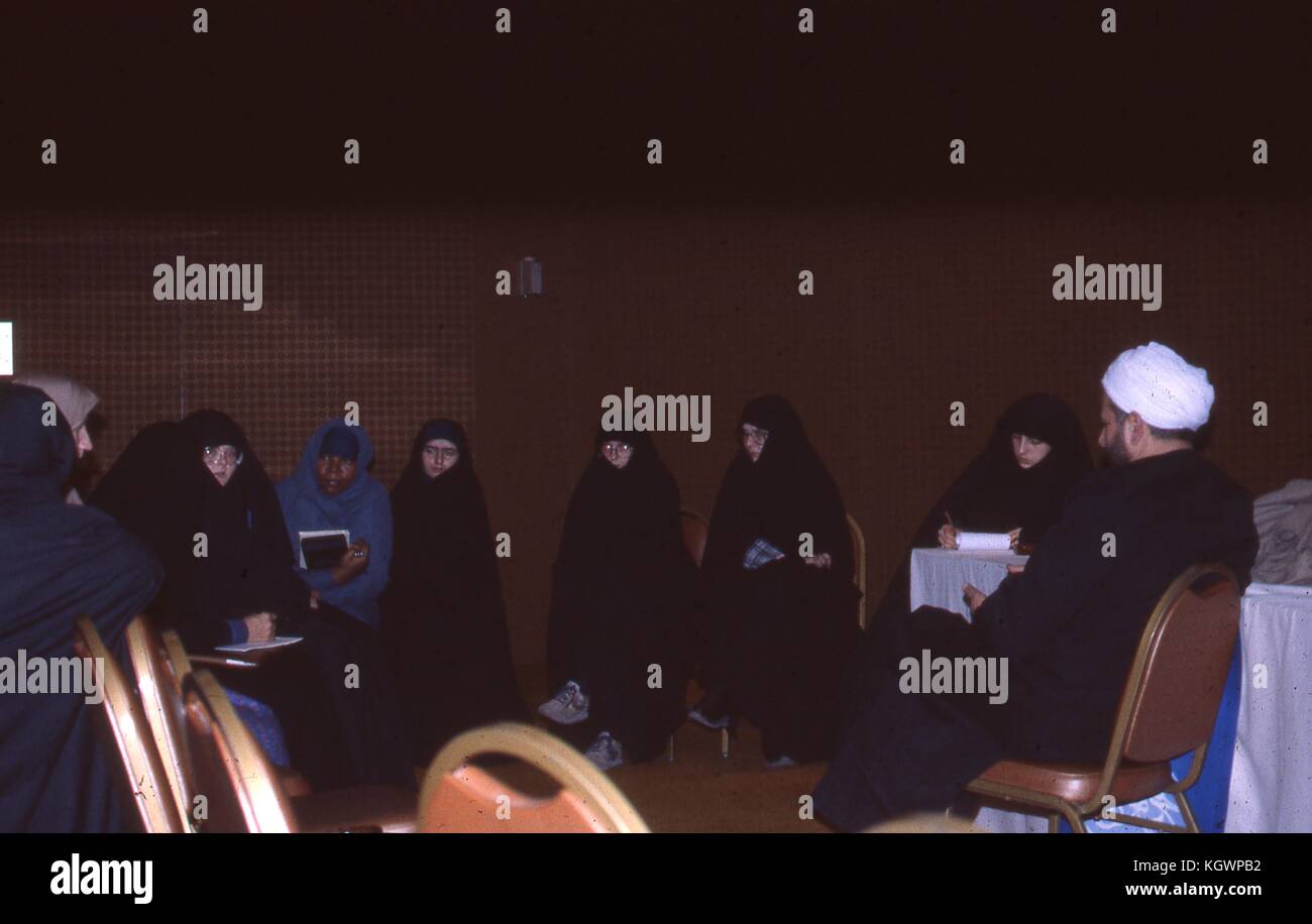 Scene of Muslim women of various ethnicities dressed in chadors, sitting in a circle taking notes, in a ballroom in Iran, March, 1983. A Muslim man in black clerical robes and a white turban sits facing the women. Extensive damage on original photo. () Stock Photo