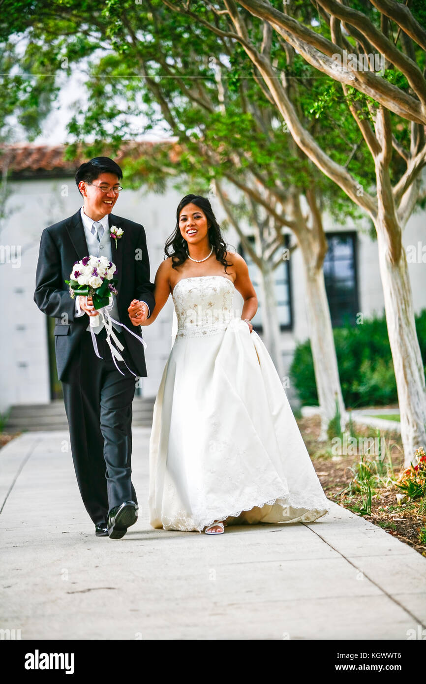 Bride and groom holding hands and walking toward the viewer outside green trees on both side of path Stock Photo