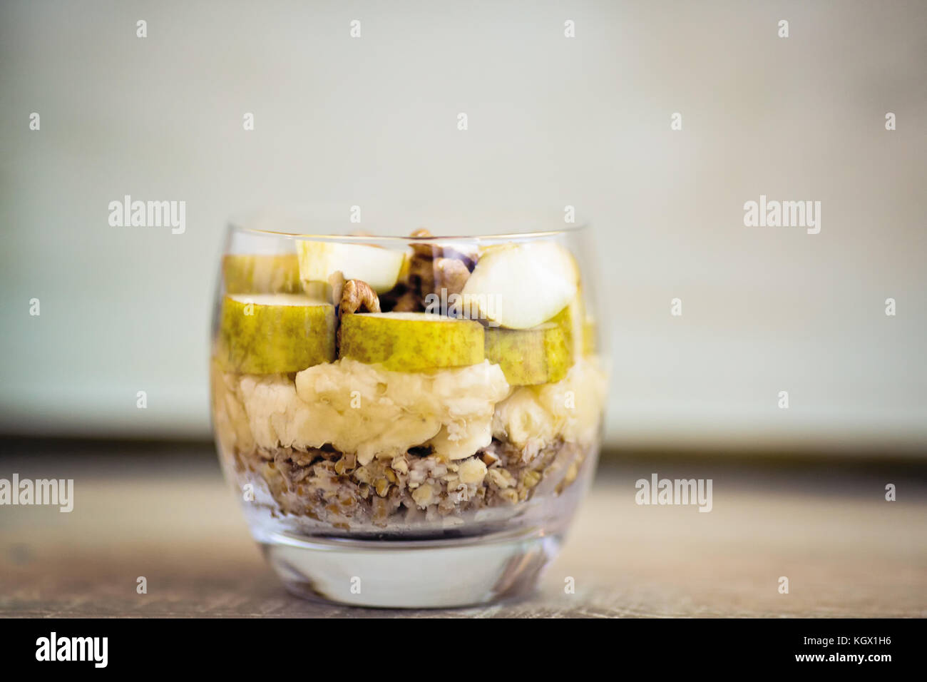 Oatmeal breakfast flavoured with mashed banana, topped with sliced pear, walnuts, sliced almonds and honey, view from side, served in a clear round gl Stock Photo
