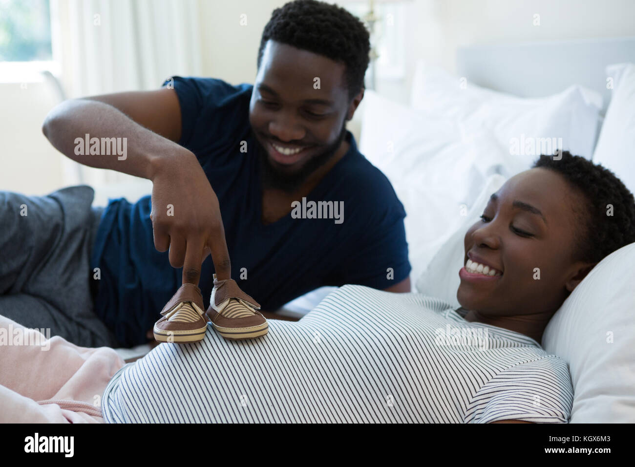 Man placing baby shoes on womans belly in bedroom at home Stock Photo