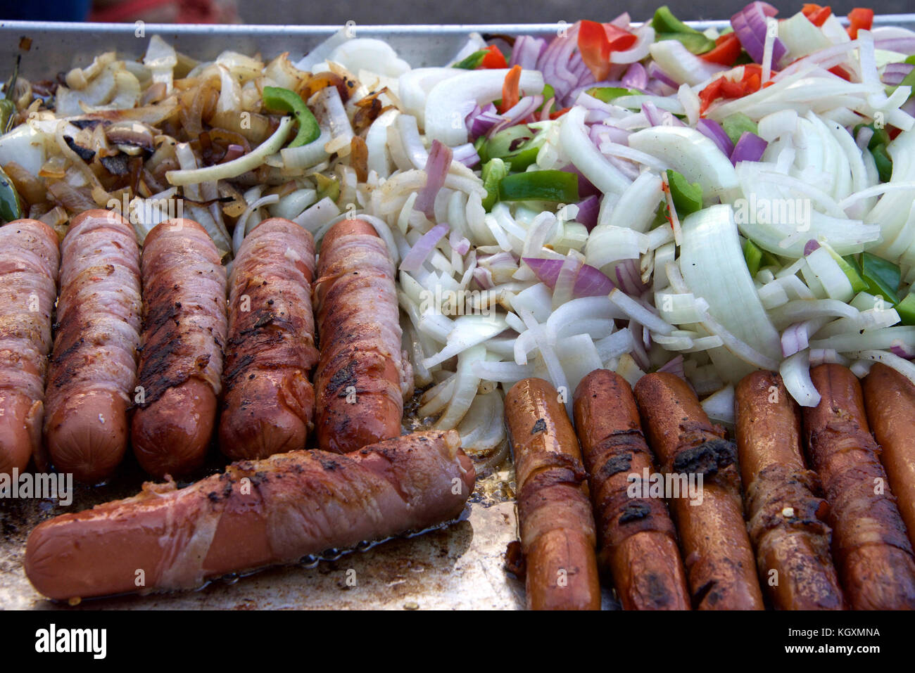 Hot dogs wrapped with thin bacon surrounded by sliced onions, bell peppers, grilling. Street