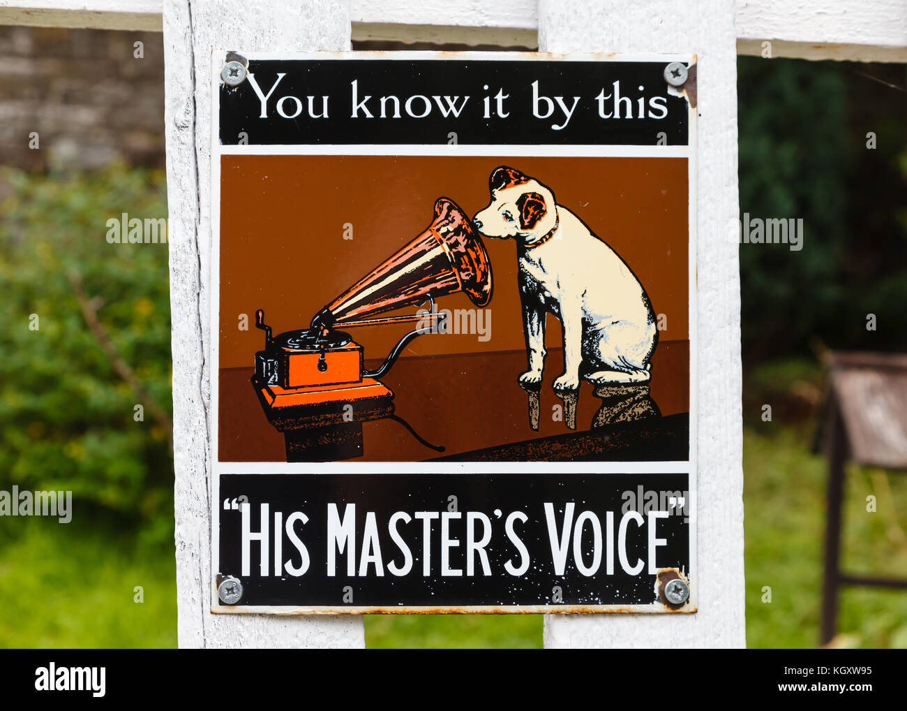 Old style tin advertising board for His Master's Voice displayed on a painted wooden fence background. Stock Photo