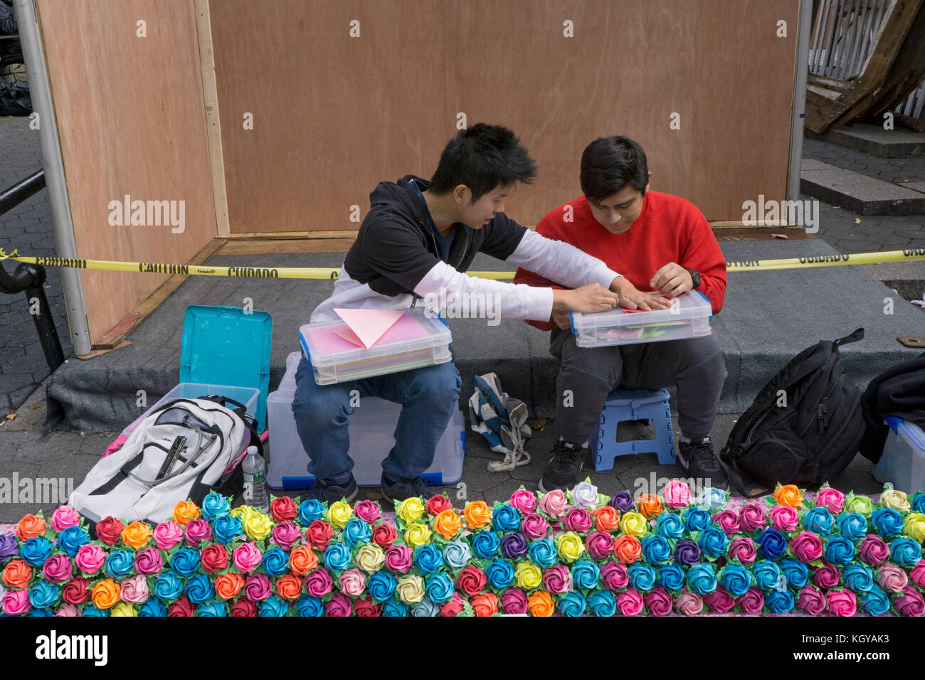 A Chinese American young man makes colorful origami flowers and instructs his friend on to do the same. At Union Square Park in New York City. Stock Photo