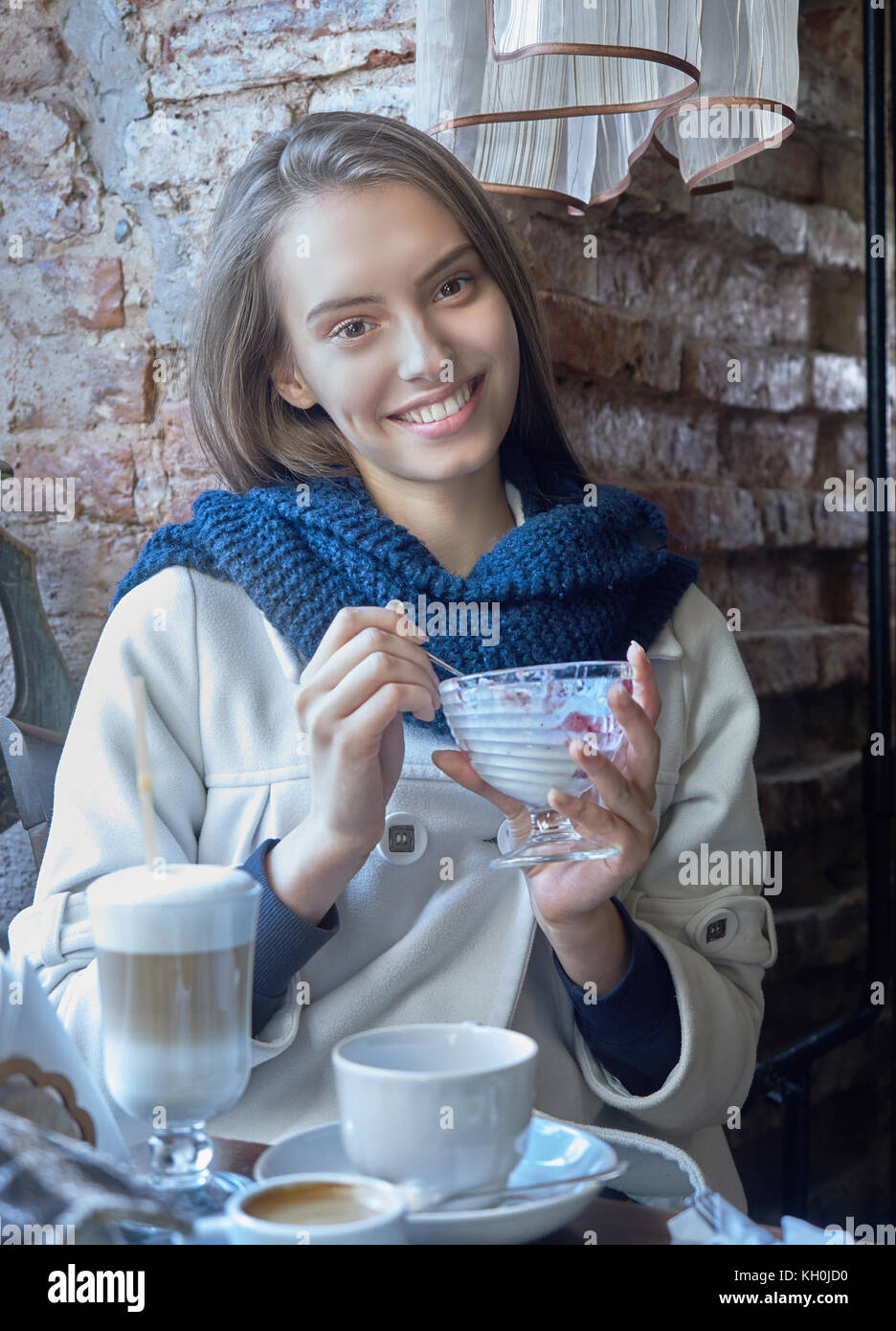 A charming young dark-haired girl is resting in a cozy cafe. Coffee is on her table. The girl enjoys the taste of ice cream. Stock Photo
