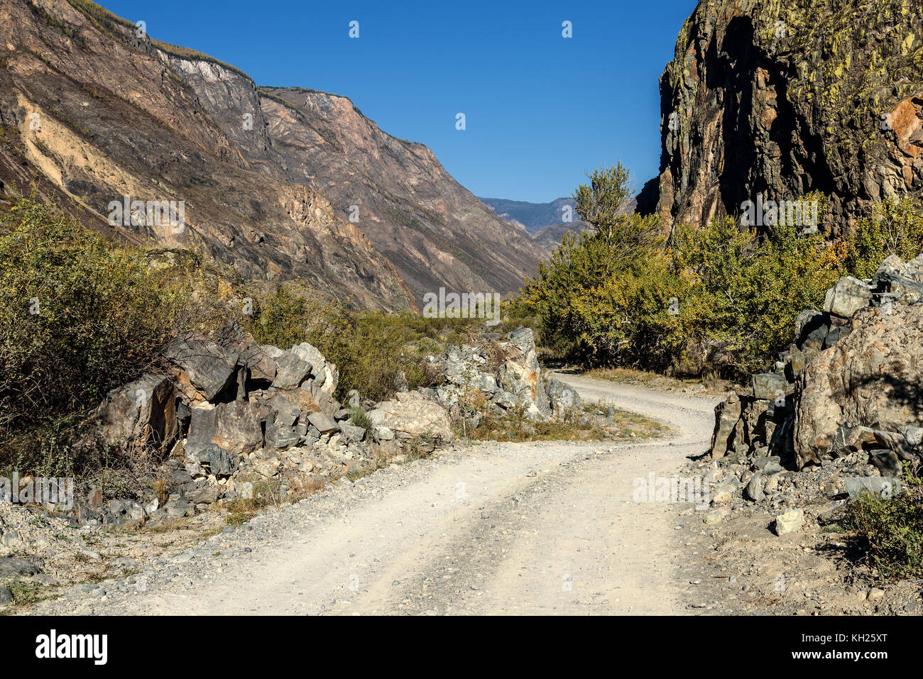 Scenic view on the gravel road with stones and vegetation on roadsides, going through the valley between the rocks on the background of blue sky Stock Photo