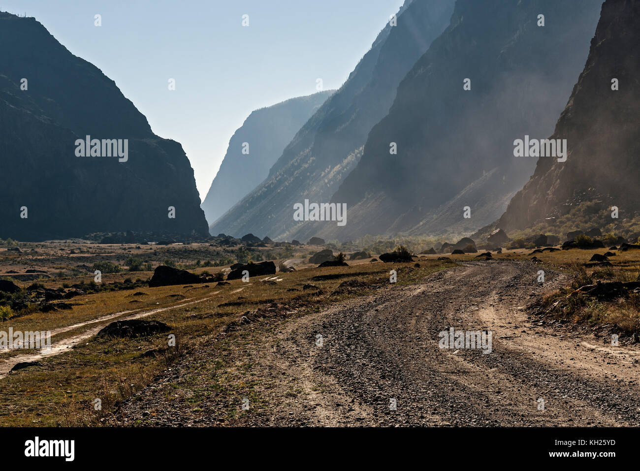 Scenic view on the gravel road with stones and vegetation on roadsides, going through the valley between the rocks on the background of sunlight Stock Photo