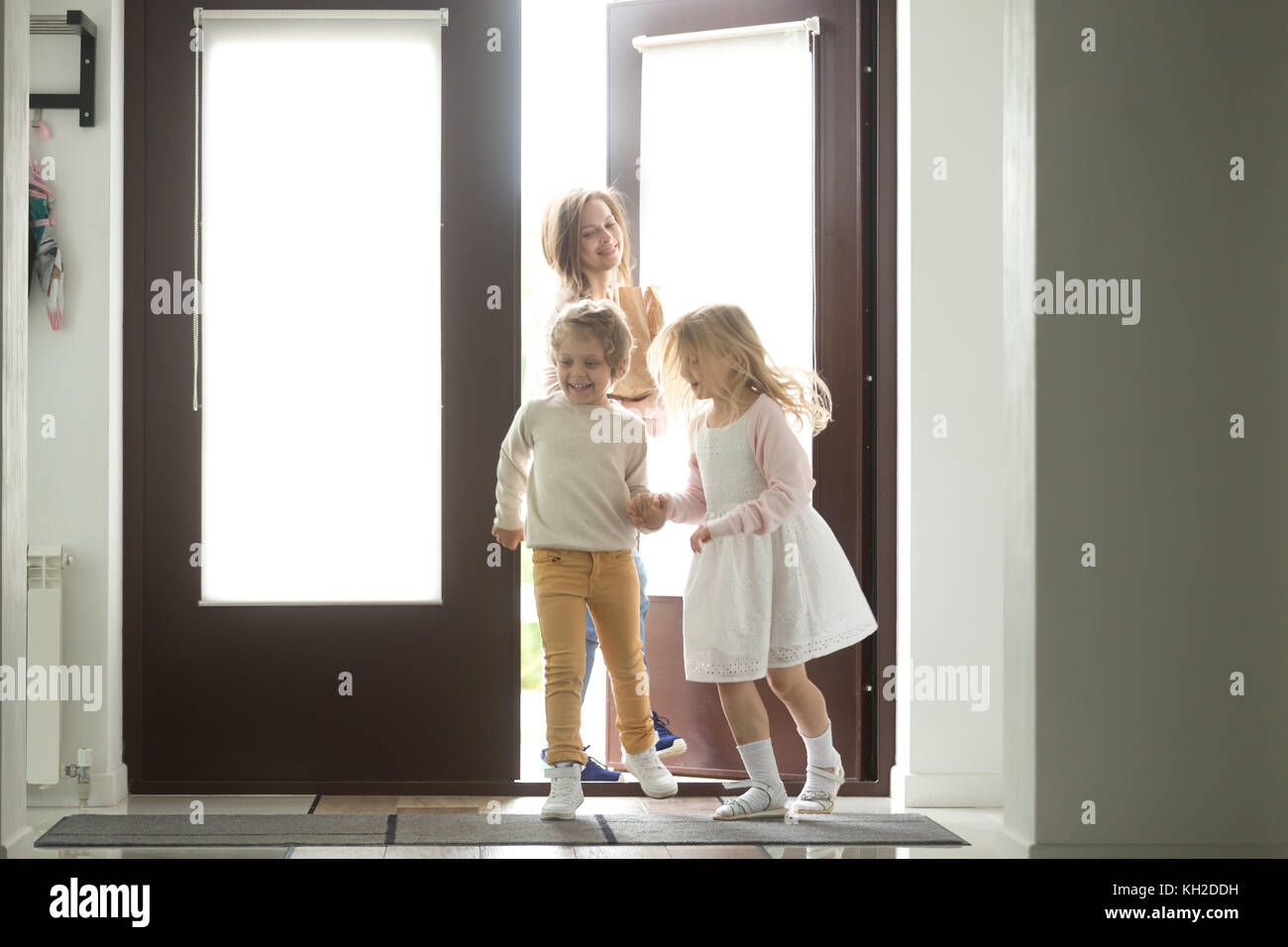 Happy young mother with son and daughter opening door coming back home from mall, single mom and small kids returning to house holding paper bag with  Stock Photo