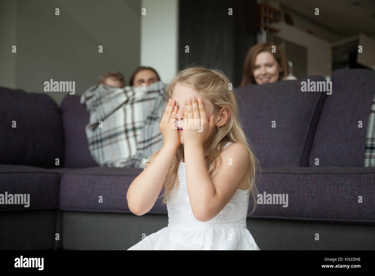 Little girl closing eyes covering face with hands playing hide and seek game with parents and brother hiding behind sofa peeking out in living room, h Stock Photo
