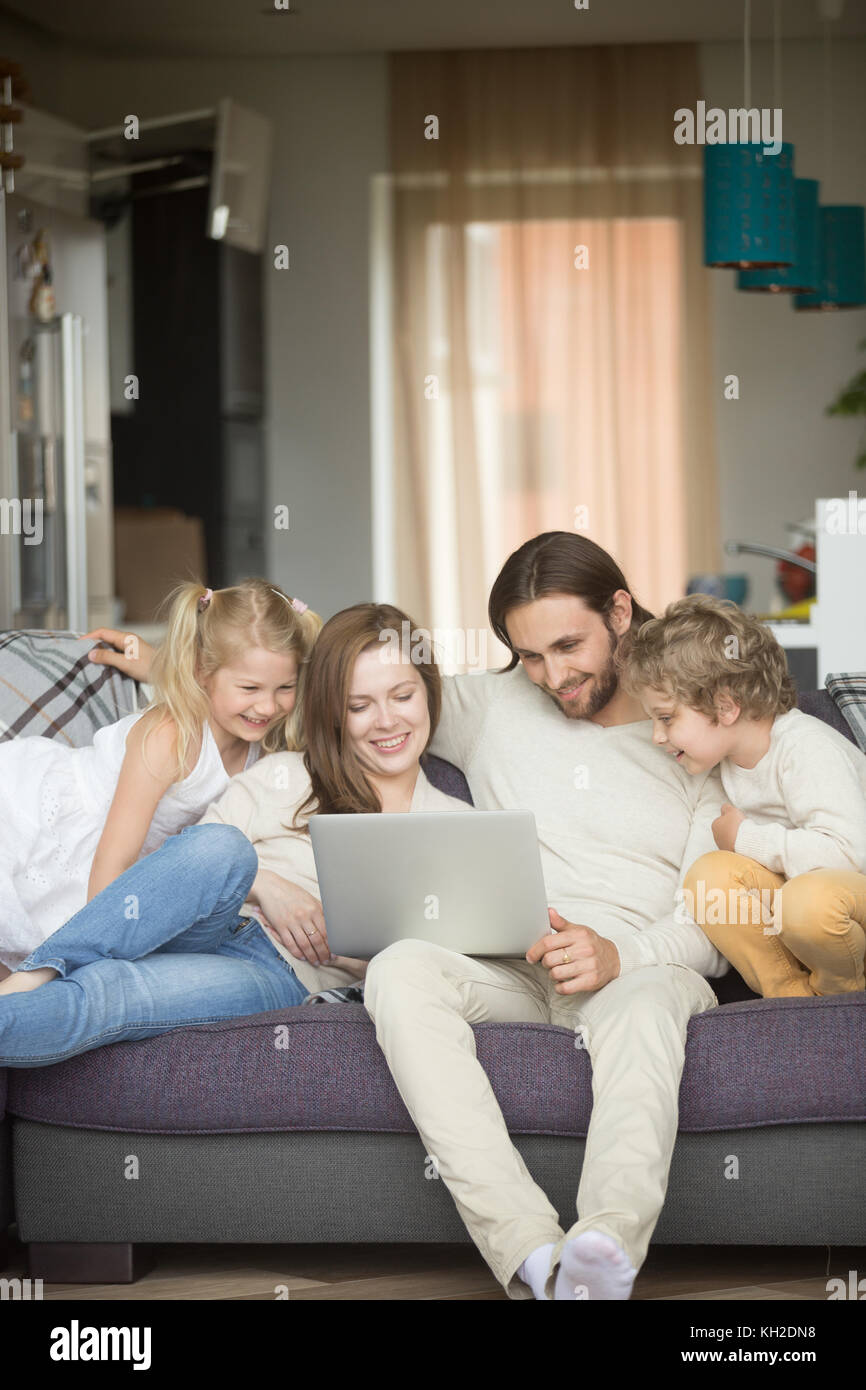 Family with children having fun using laptop sitting on sofa at home, parents and son daughter relaxing on couch holding computer, man woman with kids Stock Photo